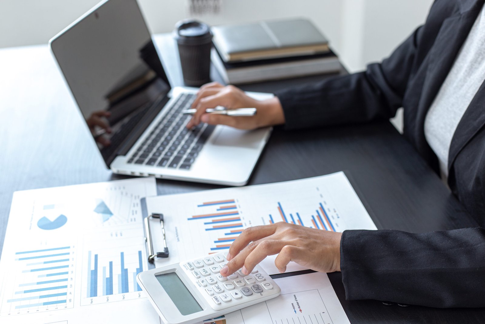 Professional using a calculator beside a laptop and financial charts